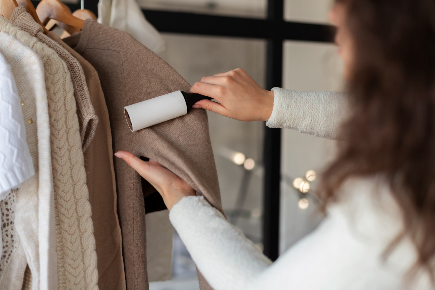 A person uses a lint roller on a brown sweater hanging on a rack, surrounded by various other clothing items.
