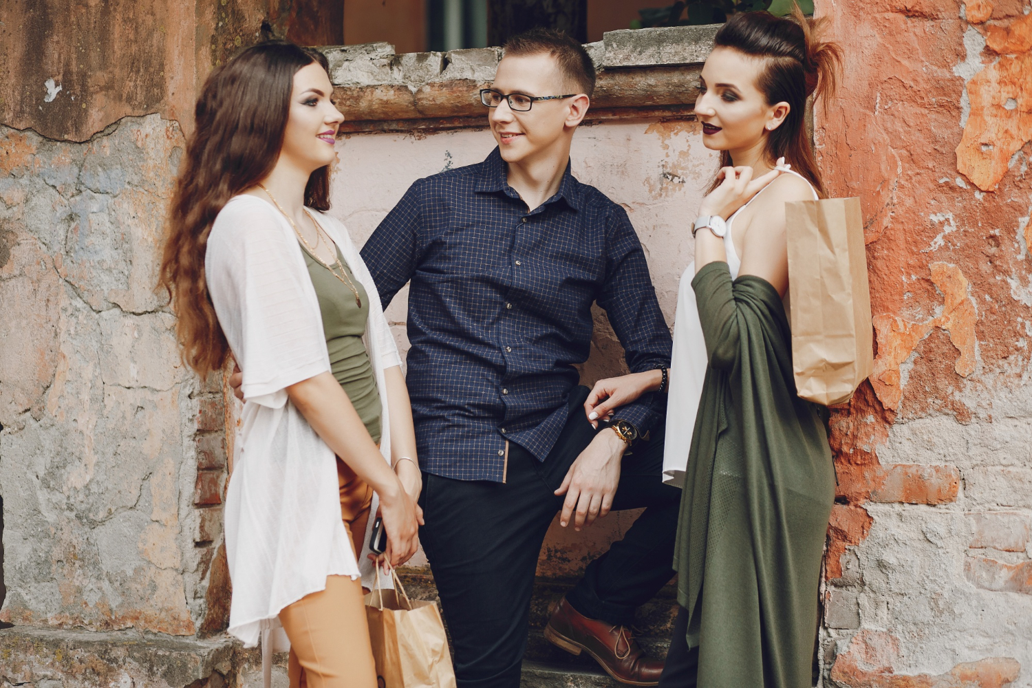 Three stylishly dressed individuals pose in front of a weathered wall, holding shopping bags, exuding a relaxed, urban vibe.