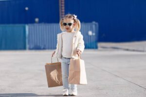 A young girl in a white sweater and jeans is walking outside, carrying two paper shopping bags, with a blue container in the background.