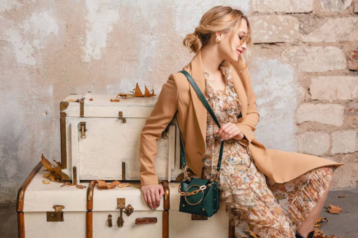 A woman in a floral dress and tan coat leans against vintage trunks, surrounded by autumn leaves on a weathered stone wall backdrop.