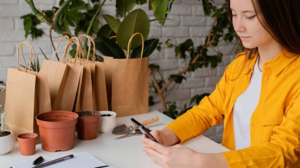 A person in a bright yellow shirt sits at a table surrounded by potted plants and paper bags, holding a smartphone.