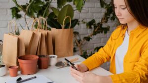 A person in a bright yellow shirt sits at a table surrounded by potted plants and paper bags, holding a smartphone.