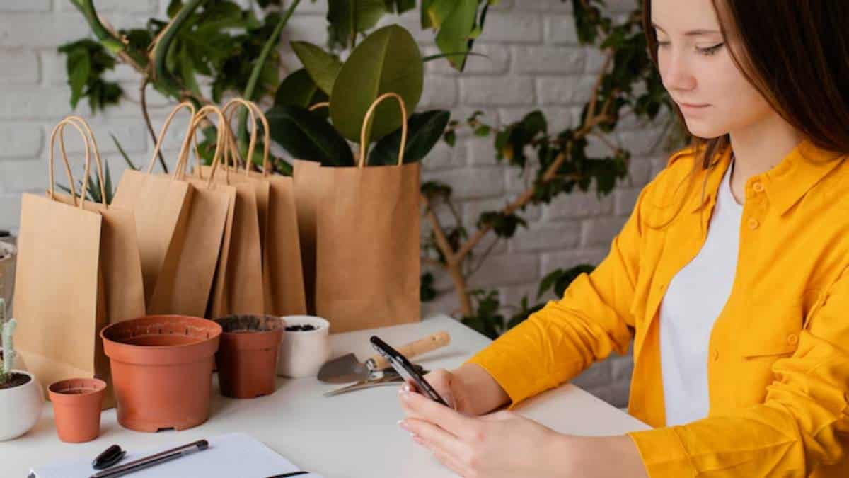 A person in a bright yellow shirt sits at a table surrounded by potted plants and paper bags, holding a smartphone.