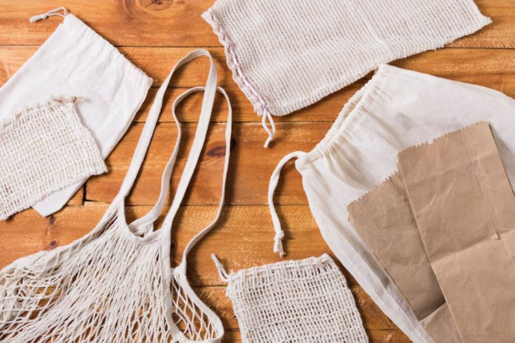 An assortment of reusable bags, including a netted tote, linen pouches, and brown paper bags, arranged on a wooden surface.