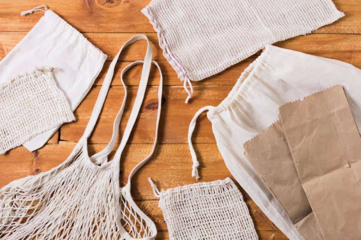 An assortment of reusable bags, including a netted tote, linen pouches, and brown paper bags, arranged on a wooden surface.