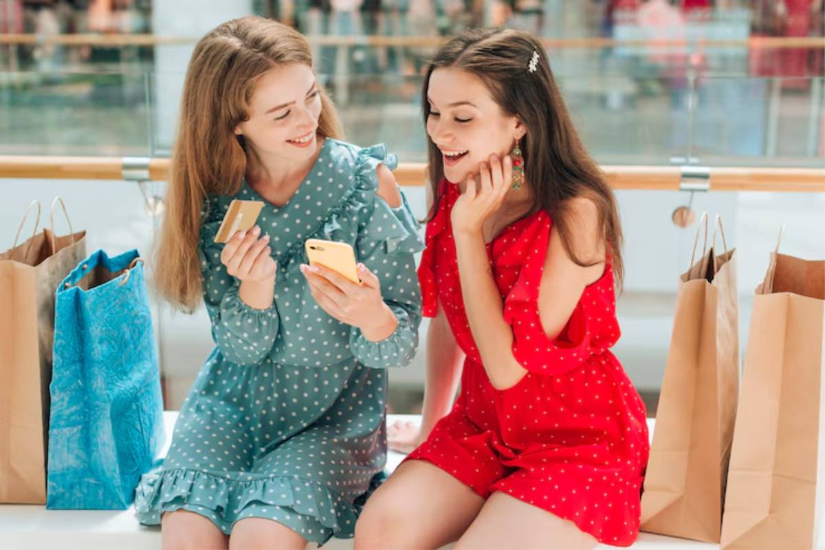 Two fashionable young women sit on a bench surrounded by shopping bags, looking at a smartphone and a credit card with interest.