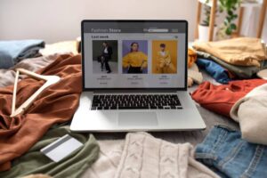A laptop displays a fashion store website on a bed surrounded by colorful clothing piles and a payment card.