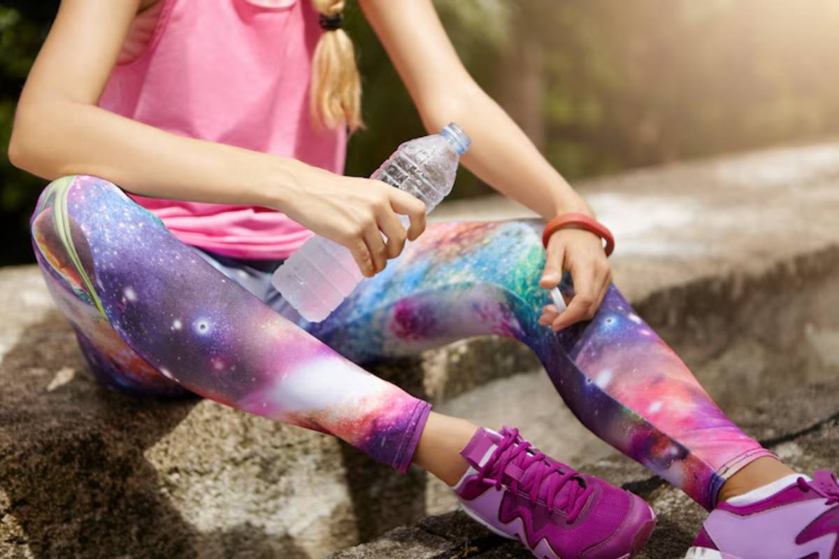 A woman in vibrant galaxy-print leggings and pink sneakers sits on a stone while holding a water bottle, looking refreshed after exercise.