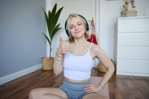 A young woman in activewear sits on a yoga mat, wearing headphones and giving a thumb's up in a well-lit room with plants.
