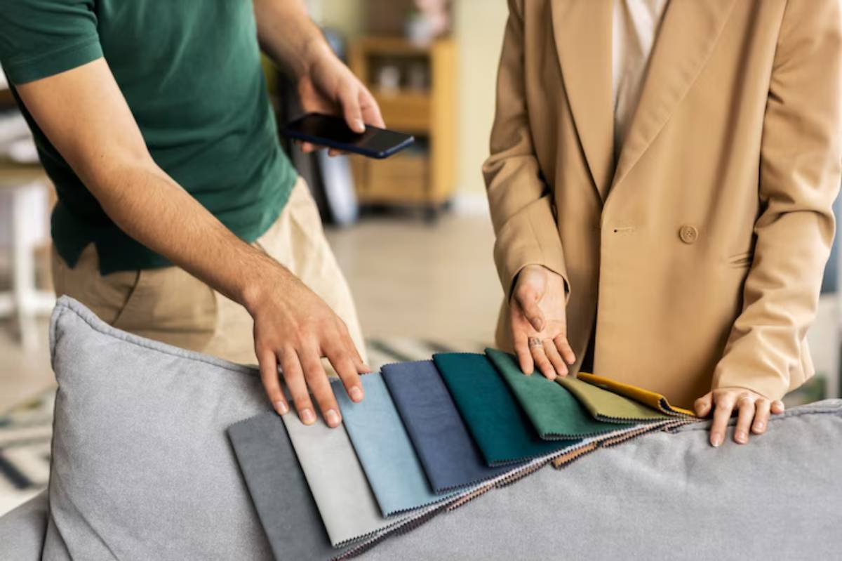 A man and woman select fabric swatches from a grey sofa, discussing color choices in a bright, cozy interior space.