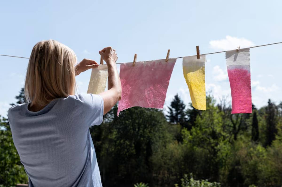 A woman hangs colorful, dyed fabric swatches on a clothesline against a sunny sky and lush green trees.