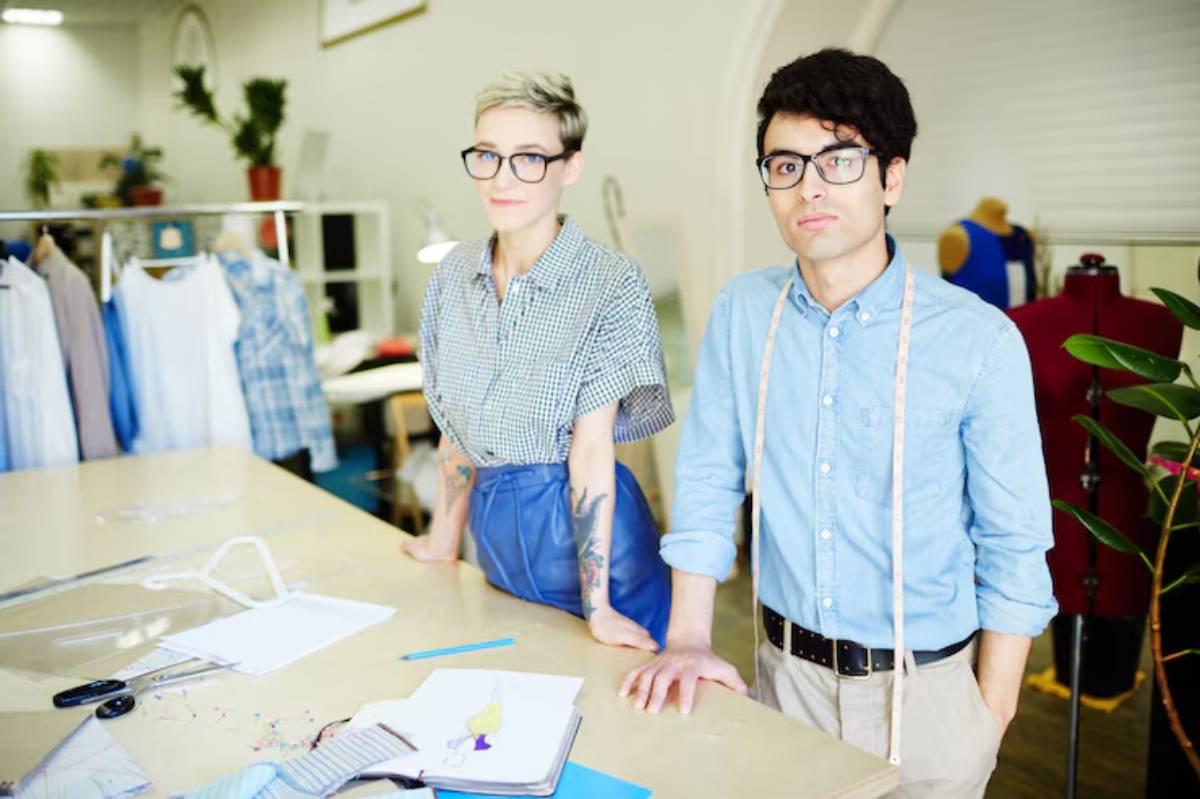 Designers stand in a bright studio surrounded by fabric, patterns, and sewing tools, engaged in a creative discussion.