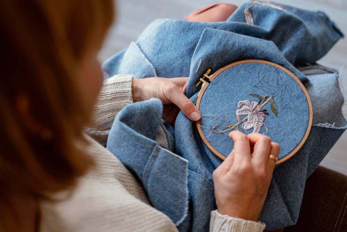 A close-up of a person embroidering a floral design on blue denim fabric, showcasing intricate stitching details.