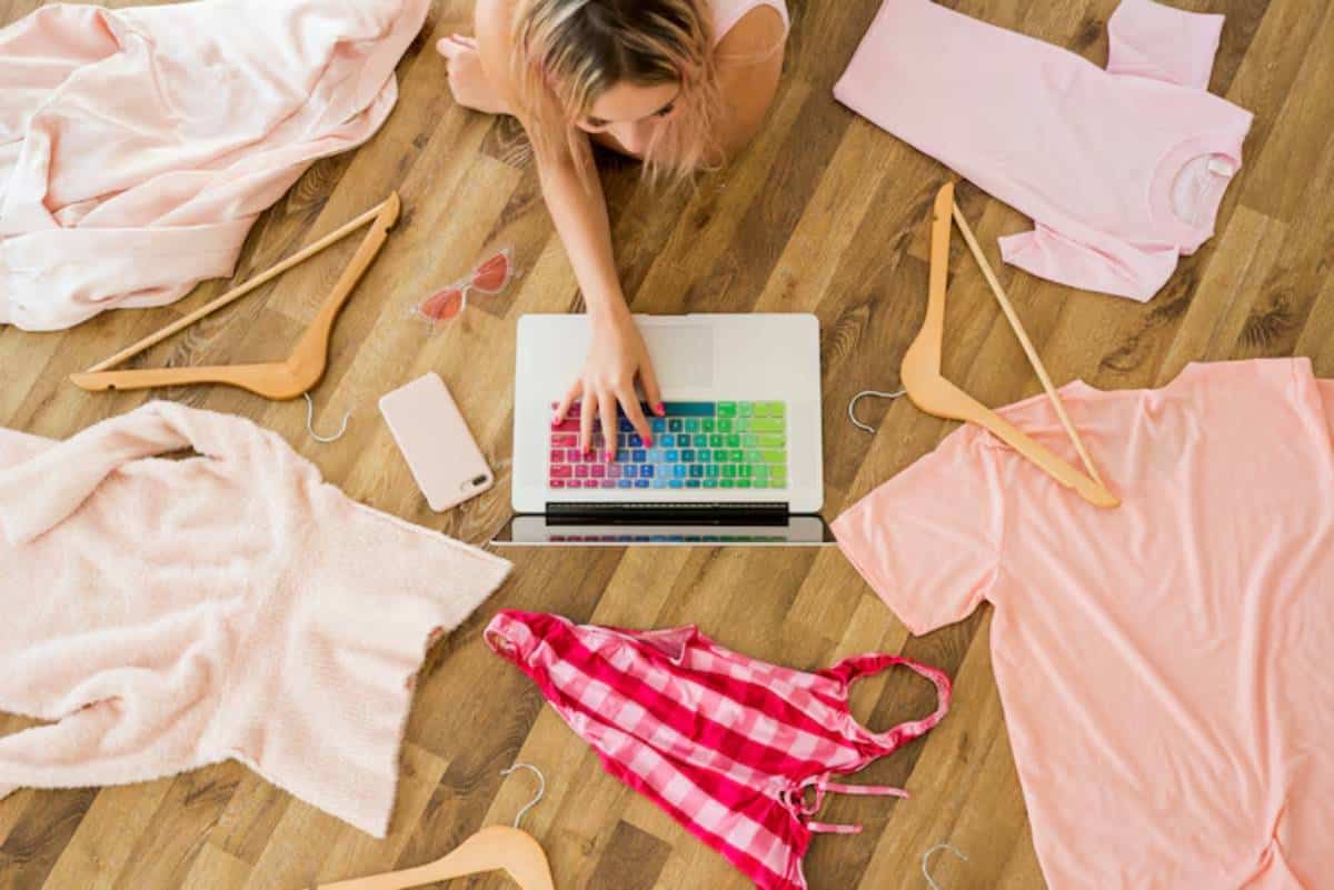 A person works on a laptop surrounded by pink clothing, hangers, and shades on a wooden floor, creating a vibrant and casual atmosphere.