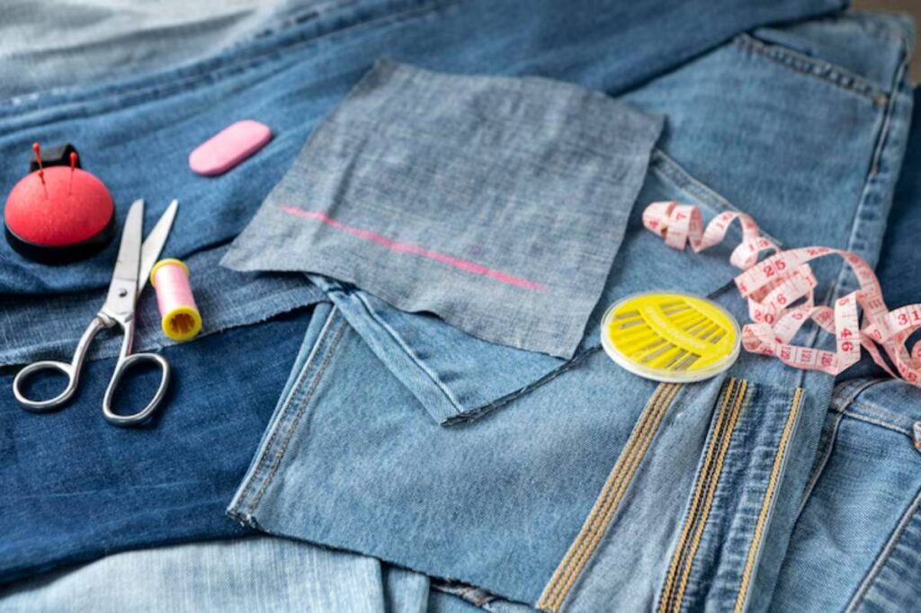 A collection of denim fabric, sewing scissors, pink and yellow thread spools, a measuring tape, and a pin cushion on a surface.