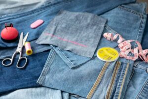 A collection of denim fabric, sewing scissors, pink and yellow thread spools, a measuring tape, and a pin cushion on a surface.