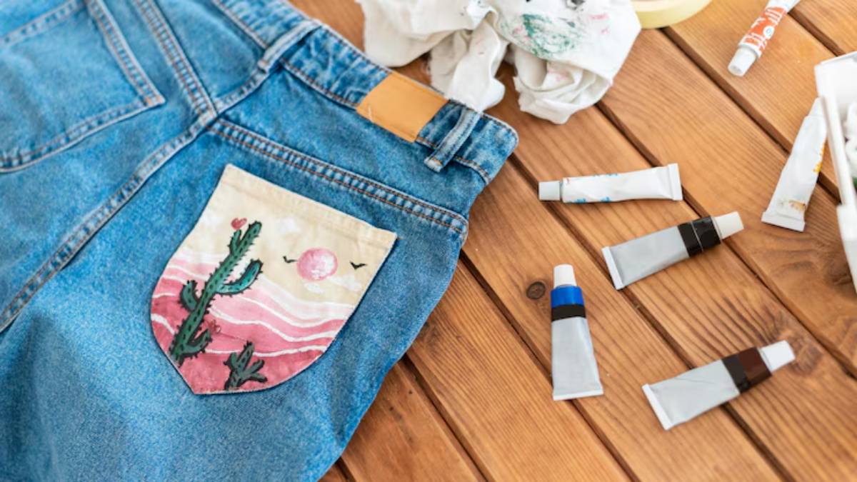 A close-up of blue jeans with a hand-painted cactus pocket, surrounded by paint tubes and a crumpled shirt on a wooden table.