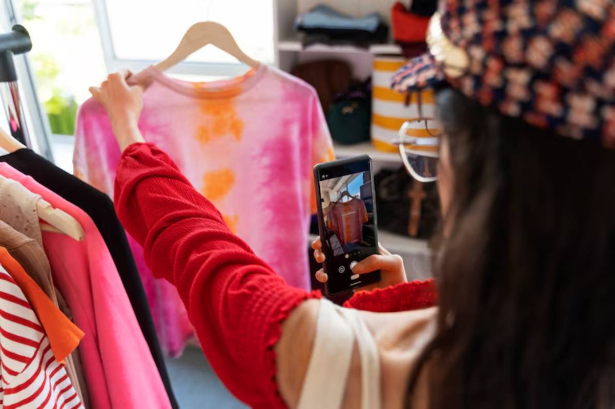 A person in a red sweater is photographing a colorful tie-dye t-shirt in a clothing store, surrounded by various garments.