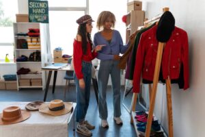 Two women browse clothing in a bright second-hand store, surrounded by hats, shoes, and a variety of colorful clothes on racks.