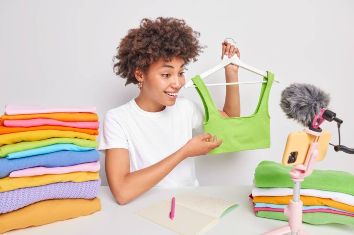 A person holds a green top, surrounded by colorful folded garments and a smartphone tripod for filming or streaming fashion content.