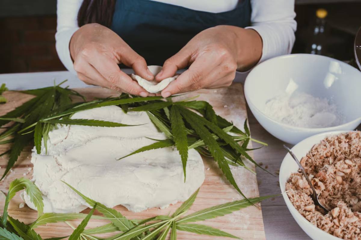 A person prepares food, molding dough with hands, surrounded by cannabis leaves, bowls of flour, and a mixture of chopped ingredients.