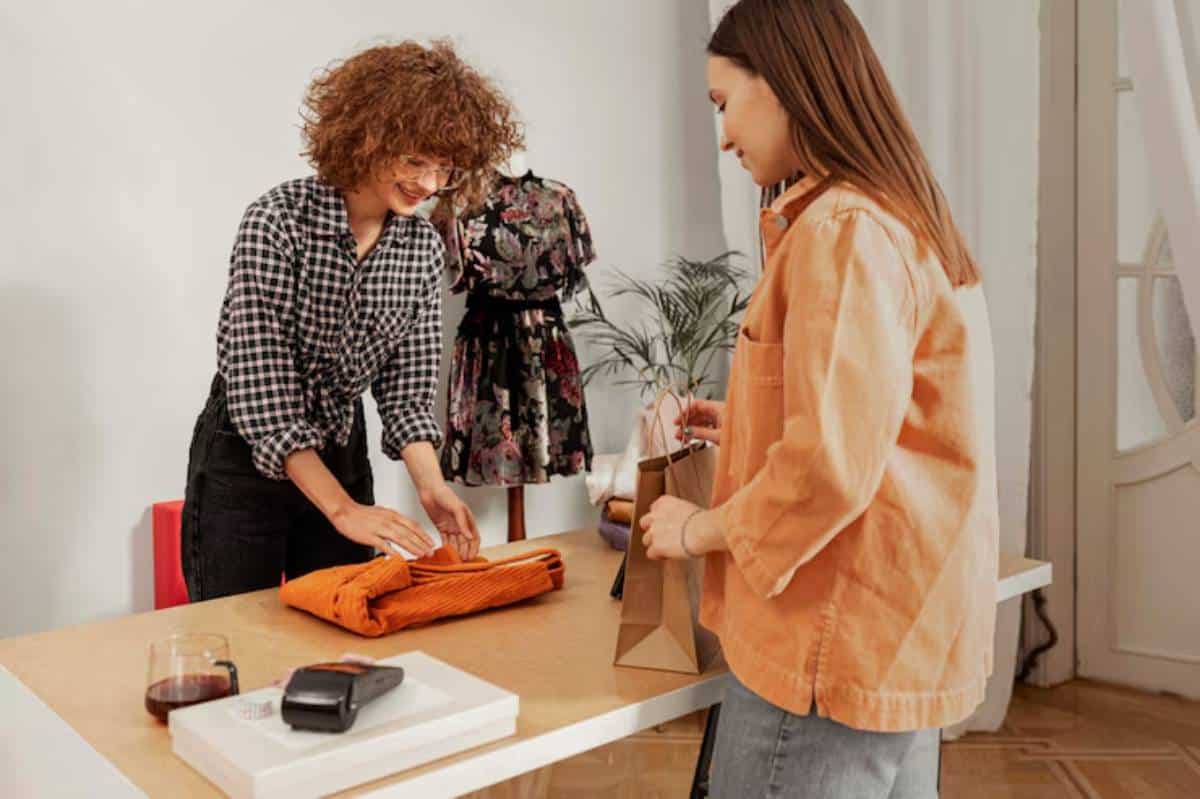 A cashier in a checkered shirt folds an orange sweater while a customer holds a brown paper bag, with a plant and dresses in the background.