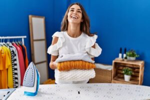 A person in a ruffled white shirt holds neatly stacked colorful sweaters, with an iron on an ironing board against a blue wall.