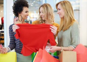 Three women are shopping together, examining a bright red shirt while surrounded by colorful shopping bags.