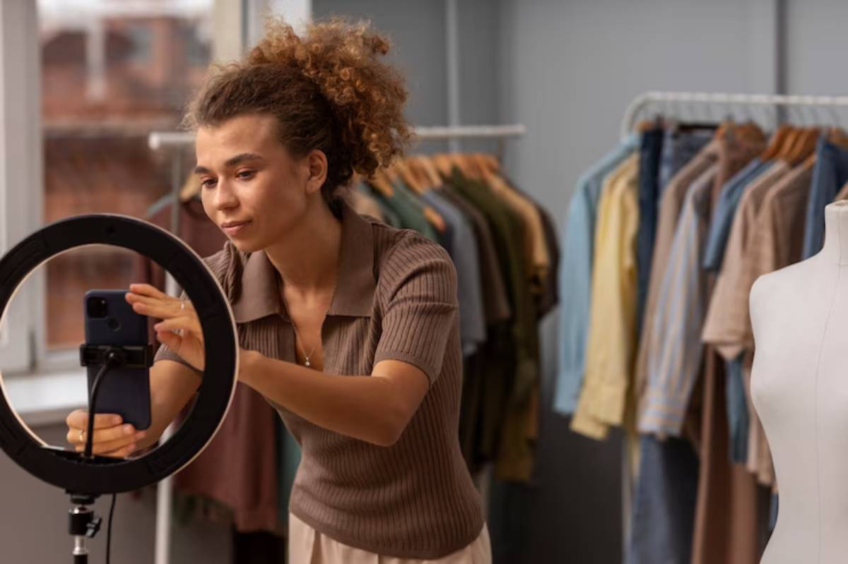 A woman with curly hair sets up her smartphone on a ring light in a clothing store, surrounded by various garments on hangers.