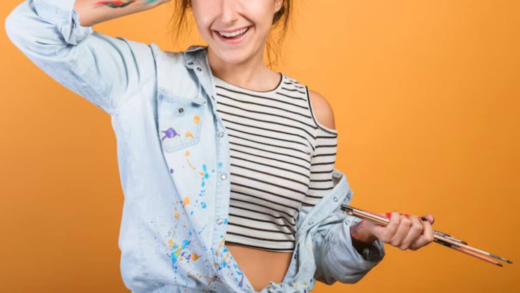 A person with a striped crop top and a denim jacket, holding paintbrushes, poses against a vibrant orange backdrop.