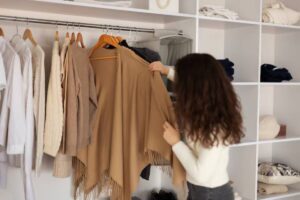 A woman with curly hair selects a tan cardigan from a neat closet, filled with various clothing items on hangers and shelves.