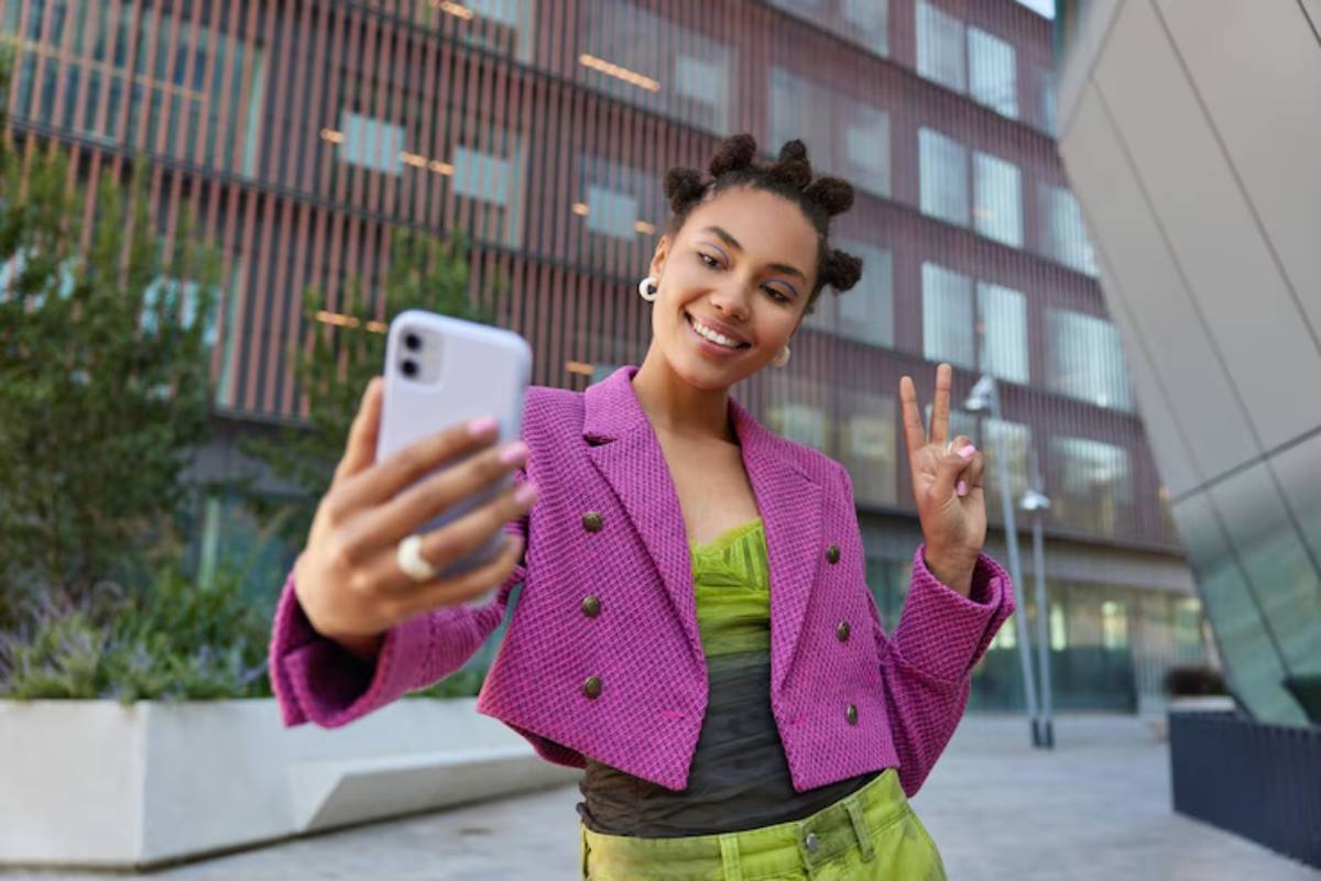 A person in a vibrant pink blazer and green outfit poses for a selfie outdoors, making a peace sign with one hand.