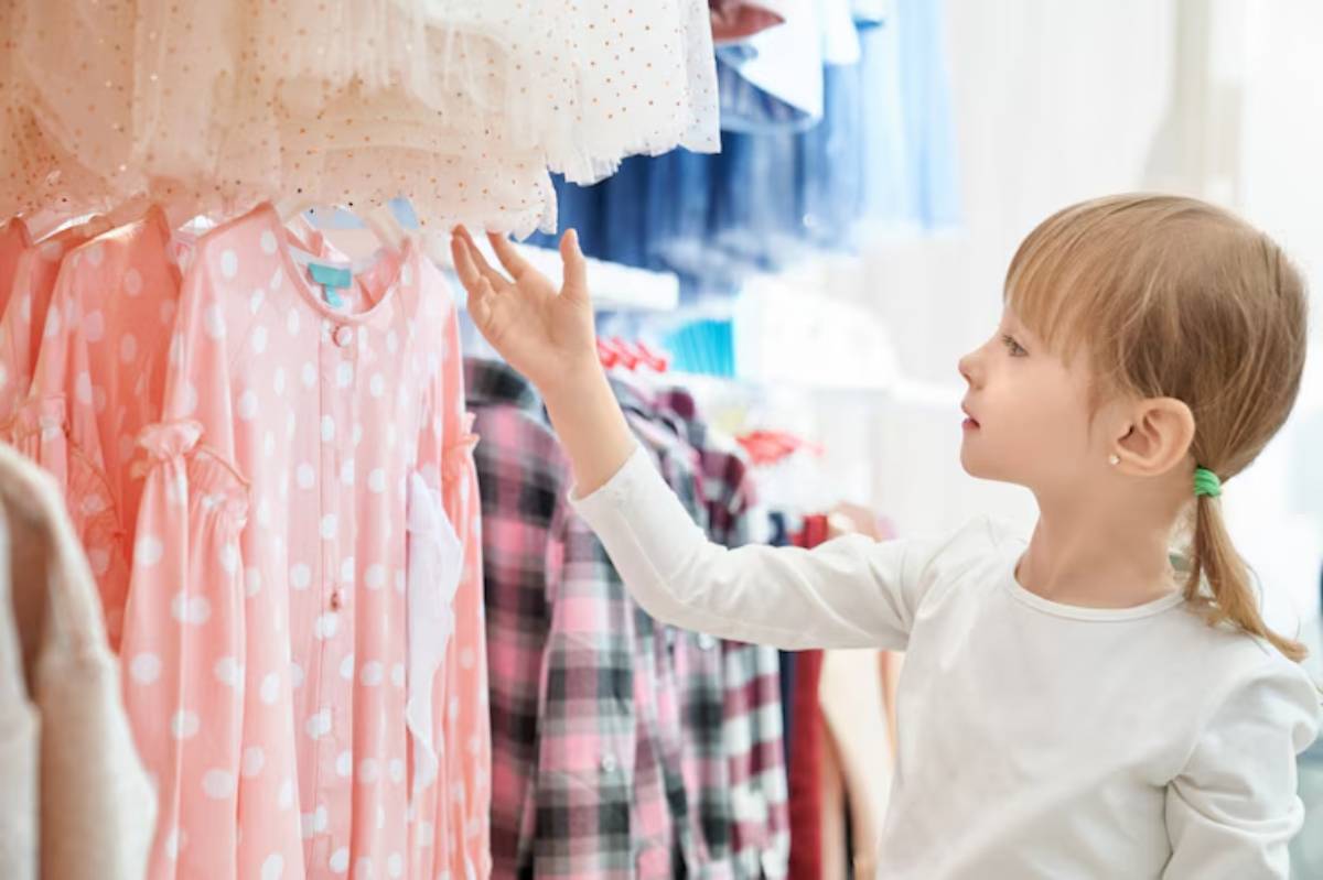 A child reaches for a pink polka dot dress on a rack in a bright clothing store, surrounded by colorful garments.