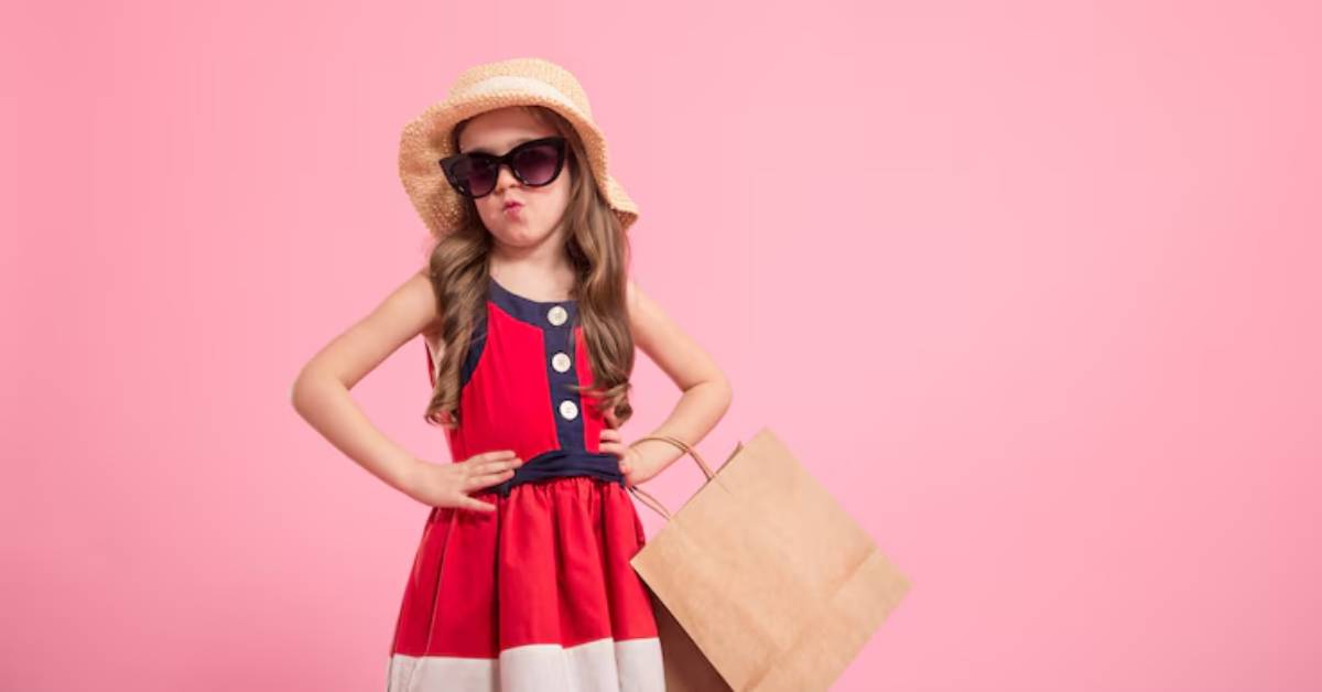 A girl in a red and white dress, wearing a straw hat, stands confidently with a shopping bag against a pink background.