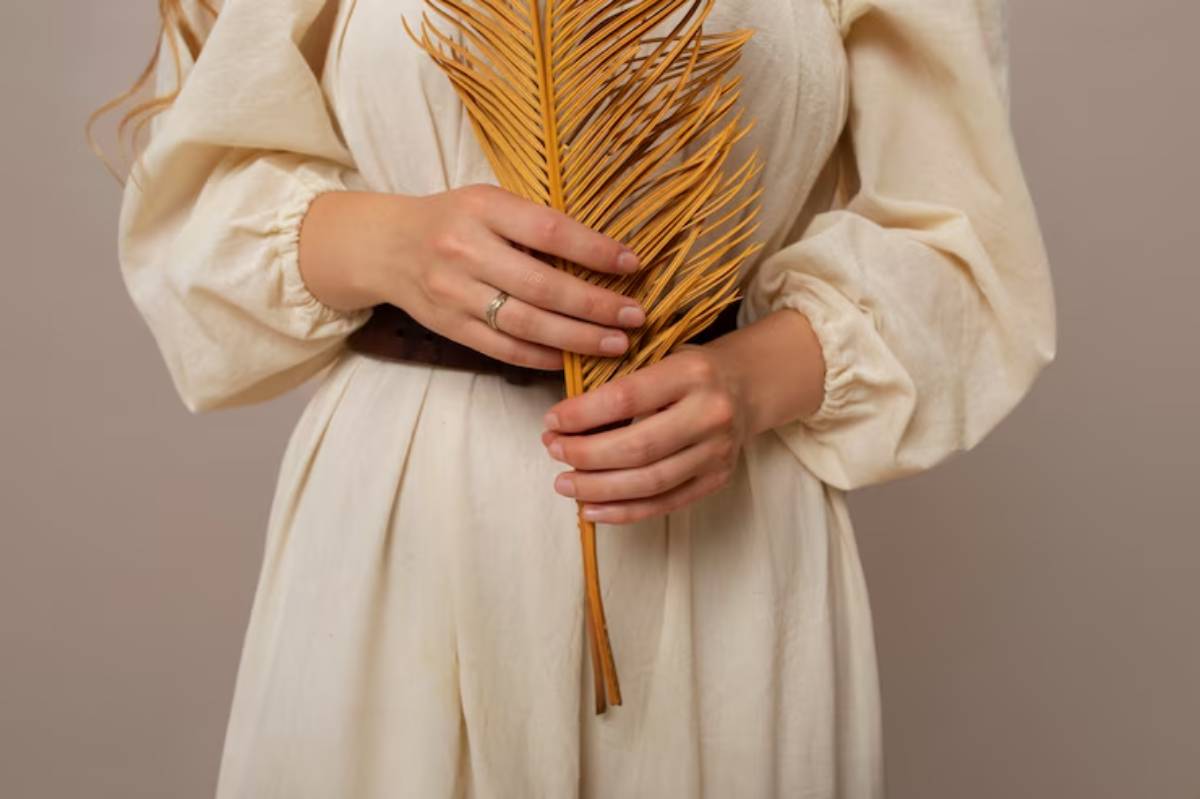 A woman in a flowing cream dress holds a dried palm leaf, showcasing her hands adorned with a simple ring against a neutral backdrop.