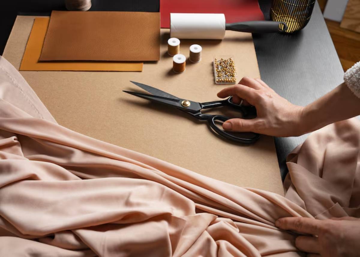 A close-up of a hand using scissors on draped fabric, surrounded by crafting materials like thread, leather, and a lint roller on a workspace.