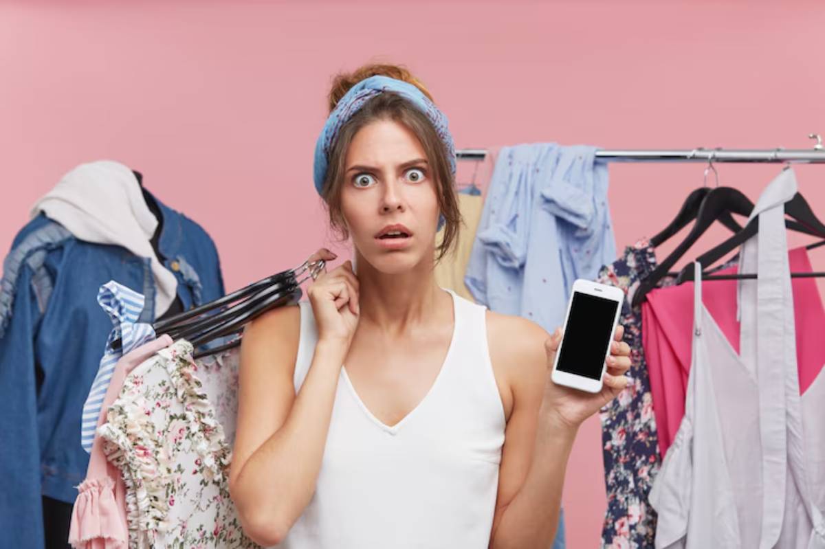 A woman holds a smartphone in one hand and colorful clothes on hangers in the other, surrounded by a pastel pink background.