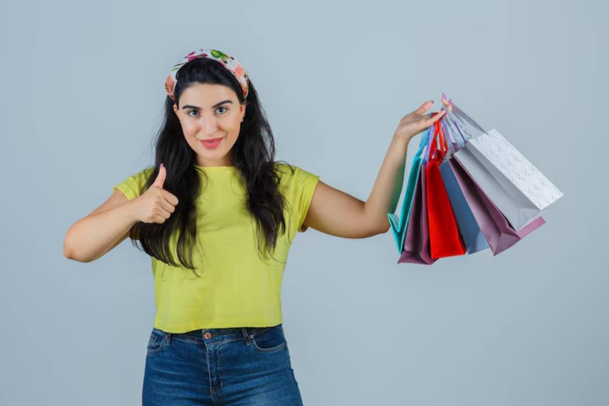 A person in a bright yellow shirt gives a thumbs up while holding several colorful shopping bags against a light gray background.