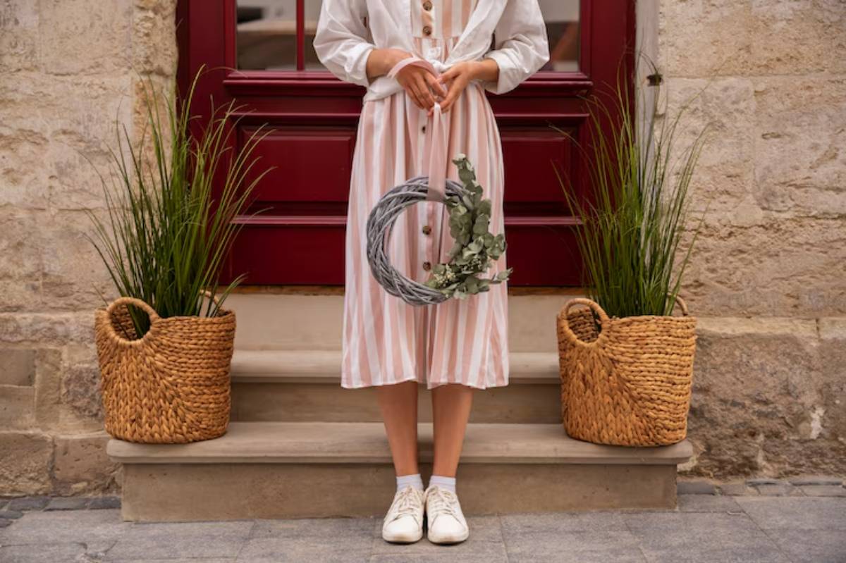 A woman in a striped dress holding a floral wreath stands on stone steps by a red door, flanked by woven baskets and greenery.