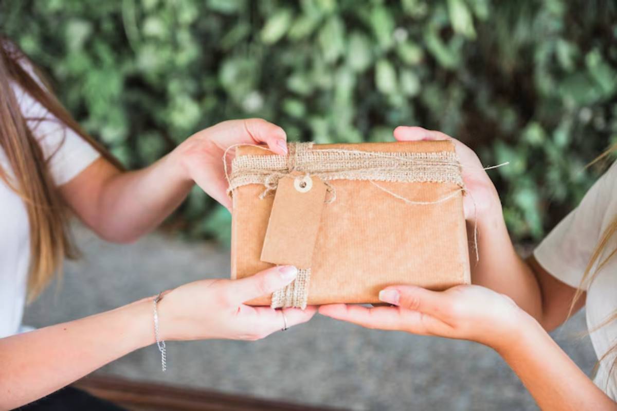 Two hands exchanging a wrapped gift with natural burlap twine and a tag, against a backdrop of greenery.