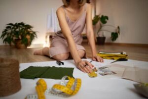 A woman in a pink dress kneels on the floor, surrounded by fabric, measuring tape, and design sketches, focusing on her sewing project.