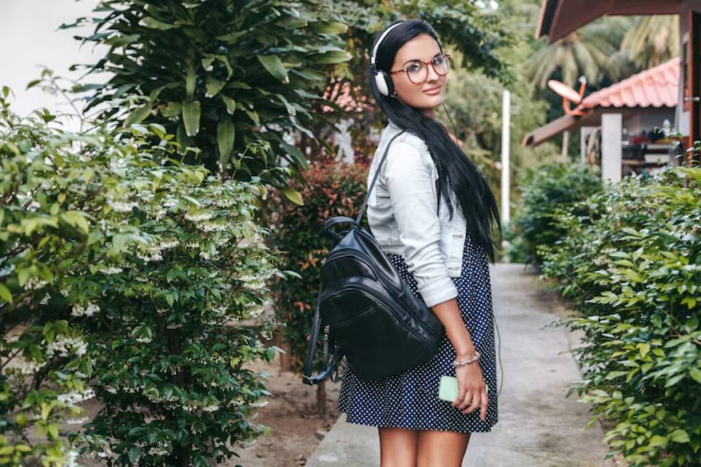 A young woman stands among lush green plants, wearing a denim jacket and polka dot dress, with headphones and a black backpack.