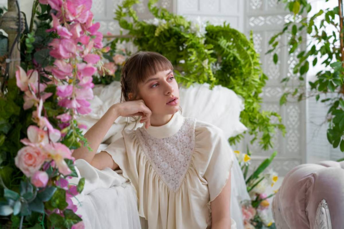 A person wearing a vintage cream-colored blouse sits gracefully among colorful flowers and lush greenery in a bright, airy setting.