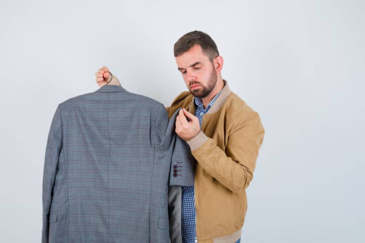 A person wearing a tan jacket holds a gray checkered suit jacket against a plain white background.