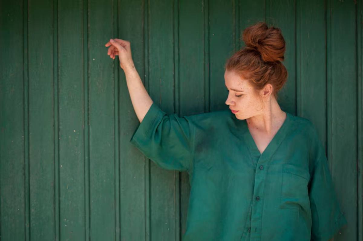 A person in a relaxed green shirt poses against a wooden green wall, with one arm lifted gracefully.