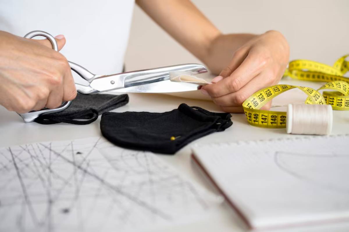 A close-up of a person cutting fabric with scissors, surrounded by sewing materials like a measuring tape, thread, and sketches.