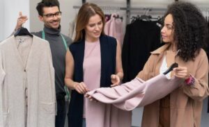 Three individuals are examining clothing items in a boutique, discussing a beige vest and a soft pink garment on hangers.