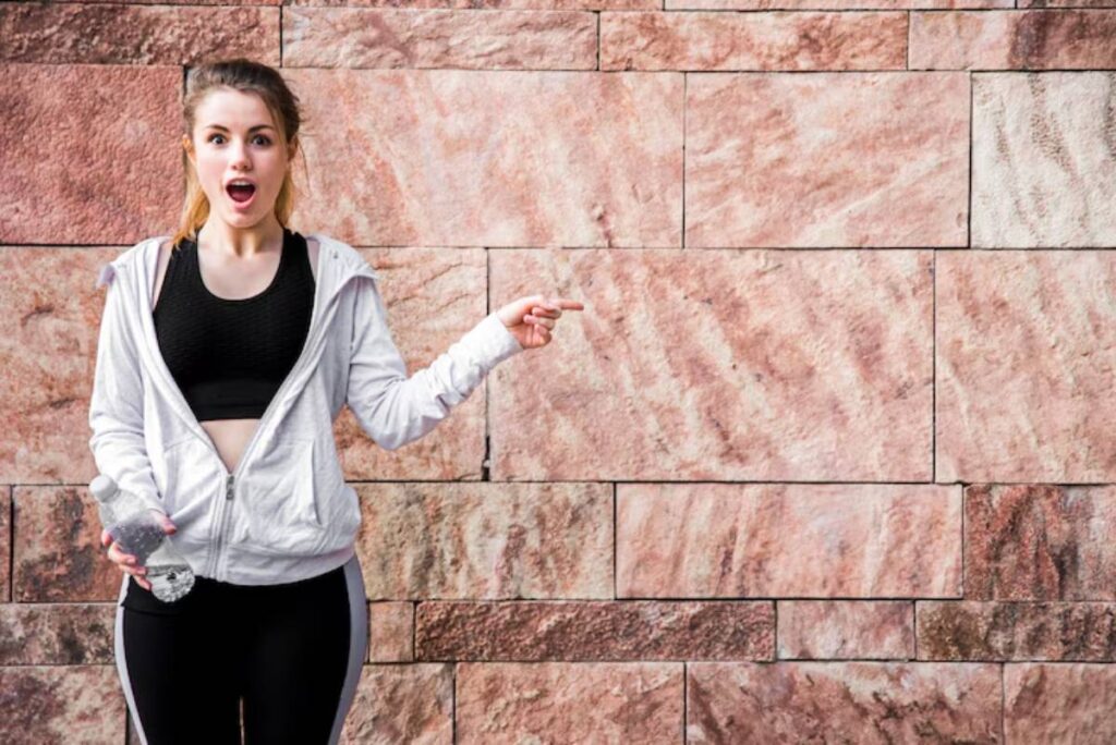 A woman in athletic wear holds a bottle of water and points, standing against a textured pink stone wall.