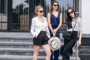 Three fashionable women stand confidently on stone steps, showcasing stylish outfits and accessories in a modern outdoor setting.