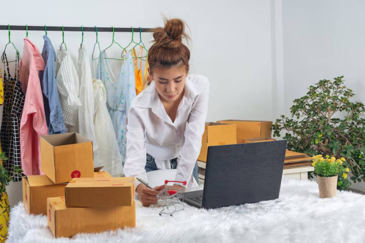 A person in a white shirt is working on a laptop surrounded by cardboard boxes and colorful clothes hanging in the background.
